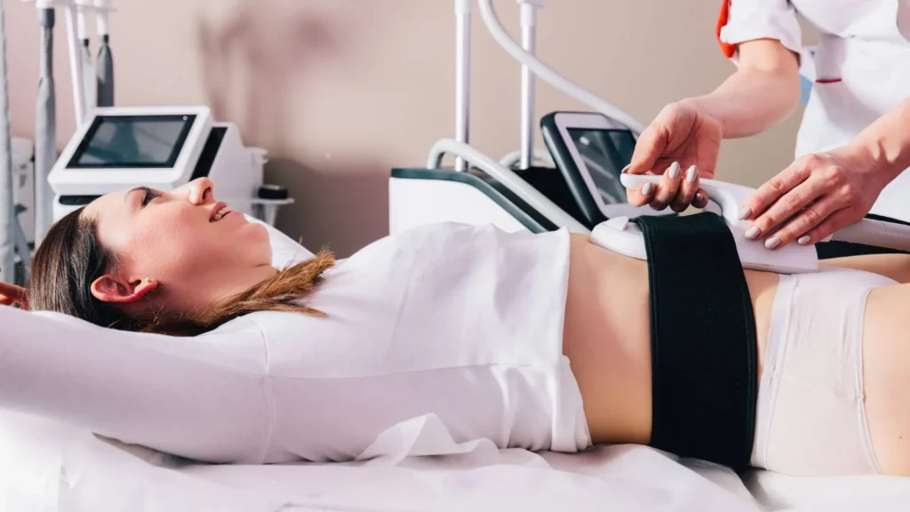 Patient lying on a treatment table while a clinician uses a handheld wand on her abdomen during a non-invasive procedure.